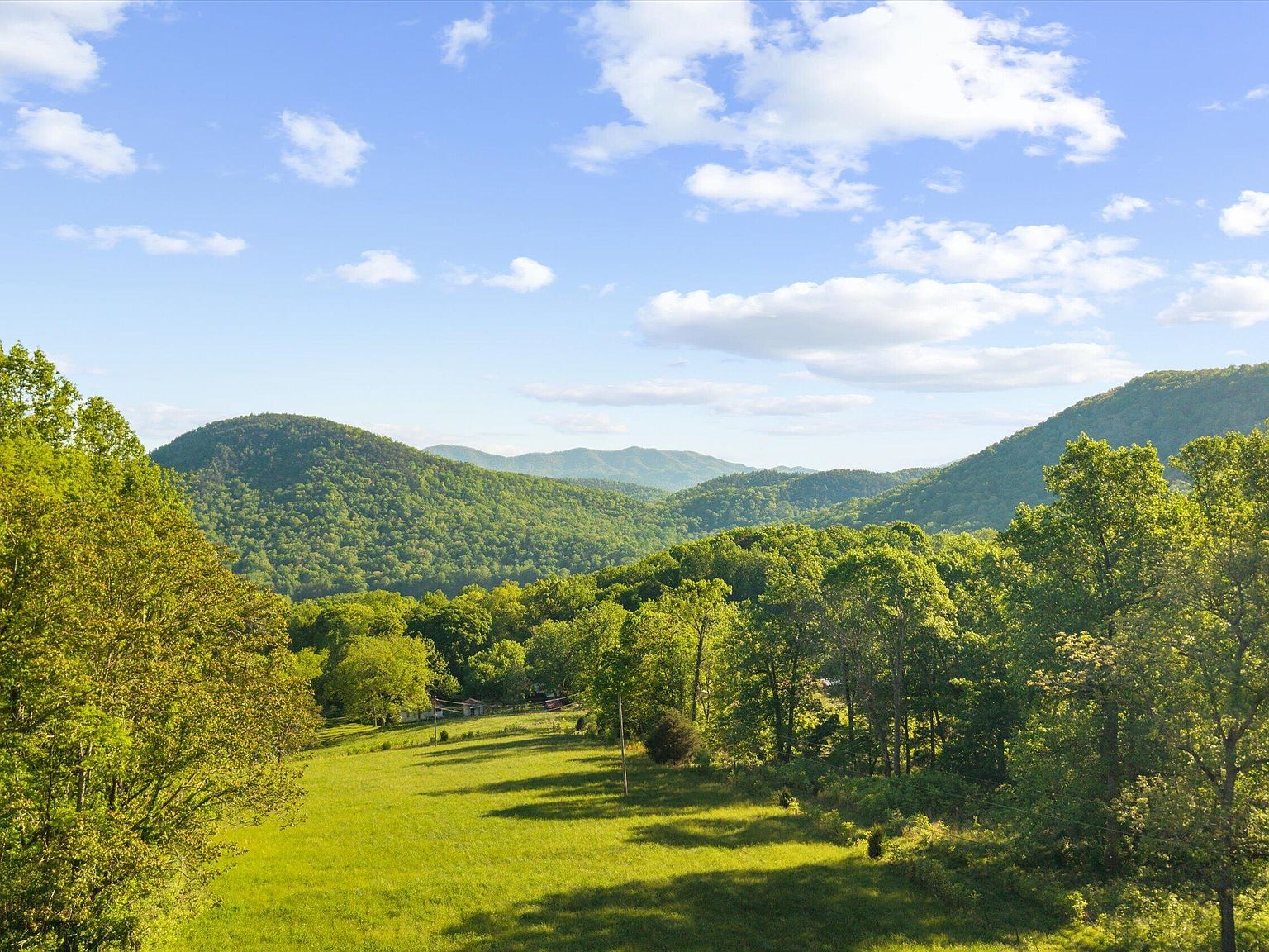 An open meadow on 172 Pam Lane framed by trees, opening to layered green ridges of the Great Smoky Mountains under a blue, cloud-dappled sky