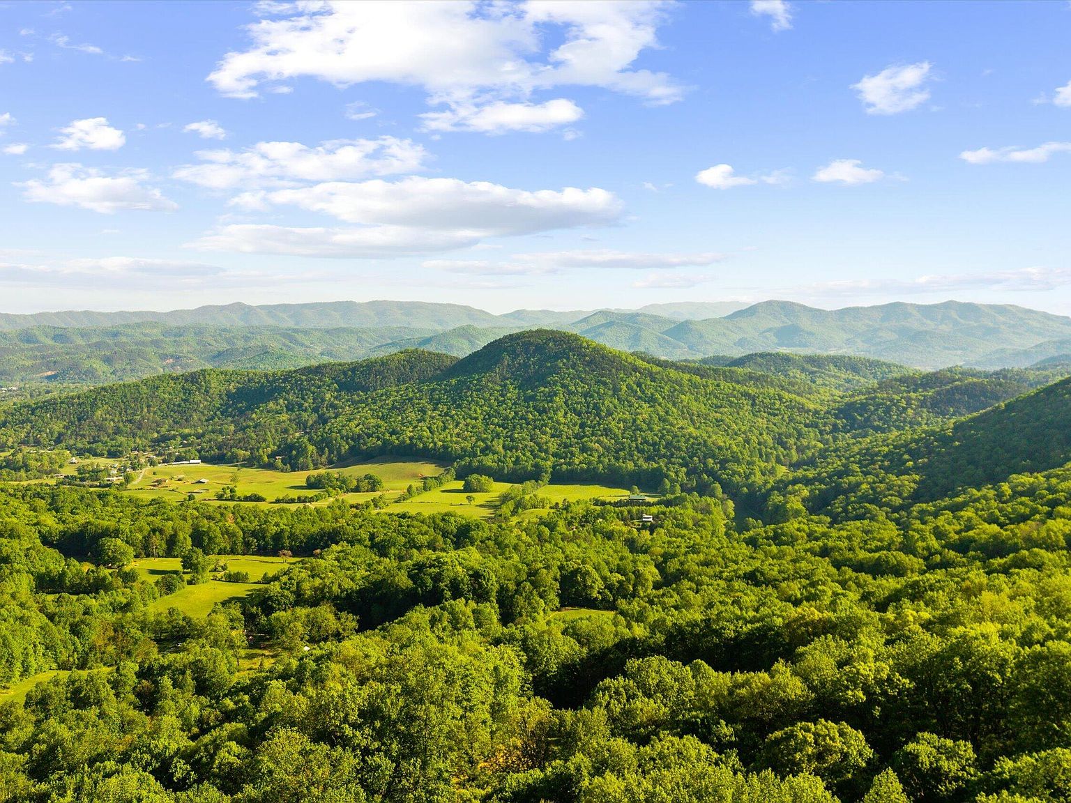Aerial view over the valley below 172 Pam Lane, with a rounded green knoll in the middle distance, cleared pastures on the valley floor, and layered ridges beyond